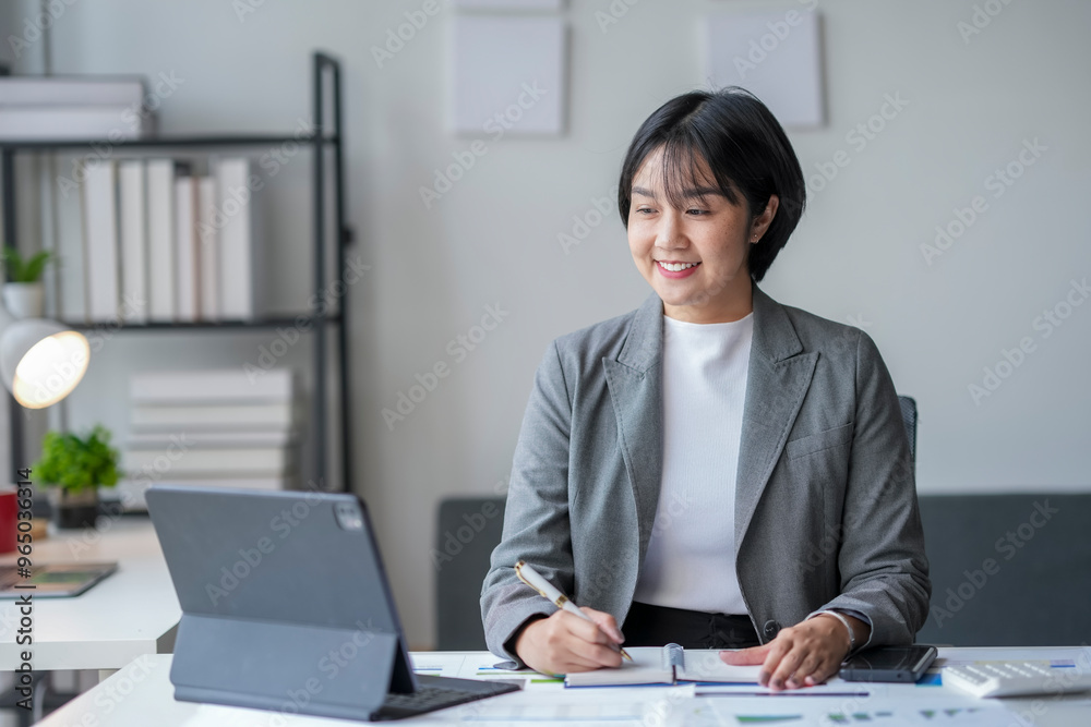 Asian businesswoman smiling and taking notes while working on a laptop