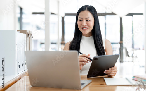 Young professional woman is focused on her digital tablet and laptop work at a bright modern office desk
