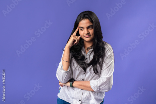 A young woman of Indian descent, wearing a white striped shirt and black smartwatch, poses thoughtfully with her finger on her temple, thought against a solid purple background