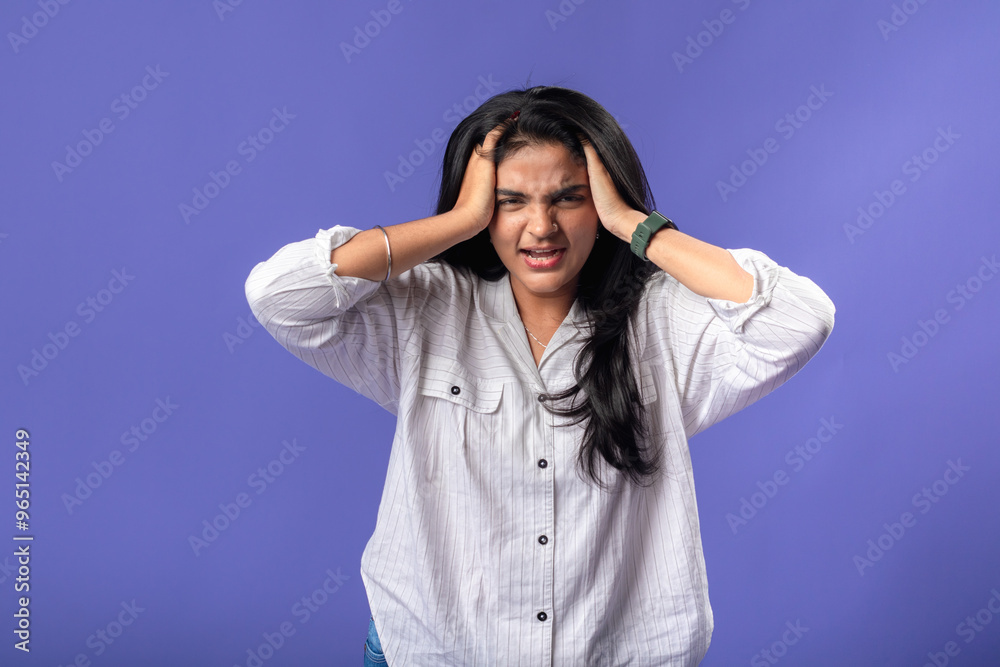 A young woman of Indian descent, wearing a white striped shirt and black smartwatch, stands against a solid purple background, expressing frustration by holding her head with both hands A young woman of Indian descent, wearing a white striped shirt and black smartwatch, stands against a solid purple background, expressing frustration by holding her head with both hands