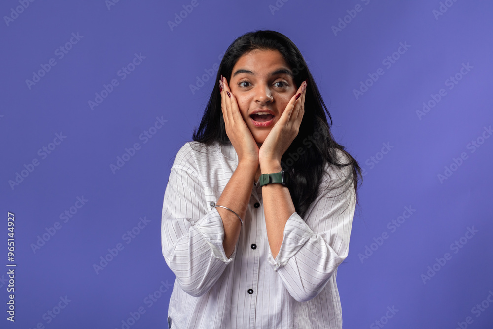 A young woman of Indian descent, wearing a white striped shirt and black smartwatch, stands against a solid purple background, with her hands on her cheeks, expressing surprise A young woman of Indian descent, wearing a white striped shirt and black smartwatch, stands against a solid purple background, with her hands on her cheeks, expressing surprise