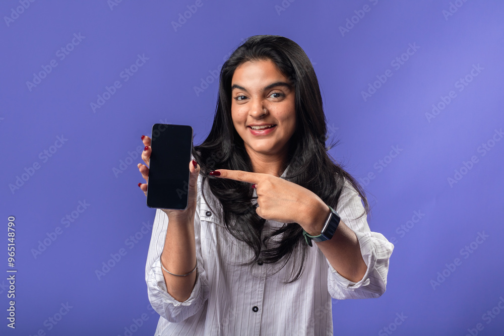 A young woman of Indian descent, wearing a white striped shirt and black smartwatch, stands against a solid purple background, smiling and extending a smartphone forward, presenting it A young woman of Indian descent, wearing a white striped shirt and black smartwatch, stands against a solid purple background, smiling and extending a smartphone forward, presenting it
