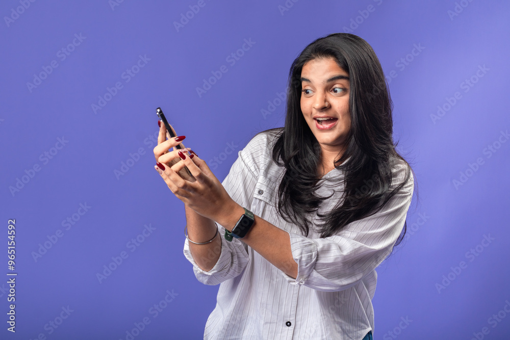 A young woman of Indian descent, wearing a white striped shirt and black smartwatch, stands against a solid purple background, smiling and holding a smartphone excitedly, as if reacting A young woman of Indian descent, wearing a white striped shirt and black smartwatch, stands against a solid purple background, smiling and holding a smartphone excitedly, as if reacting