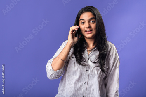 A young woman of Indian, wearing a white striped shirt and black smartwatch, against a solid purple background, holding a smartphone to her ear, as if having a phone conversation.