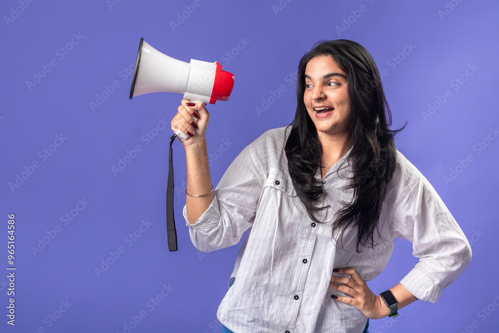 A young woman of Indian descent, wearing a white striped shirt and black smartwatch, stands against a solid purple background, smiling and holding a megaphone, suggesting communication. A young woman of Indian descent, wearing a white striped shirt and black smartwatch, stands against a solid purple background, smiling and holding a megaphone, suggesting communication.