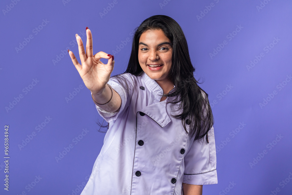 A young woman of Indian descent, dressed in a light purple chef's coat, stands against a solid purple background, smiling and making an A young woman of Indian descent, dressed in a light purple chef's coat, stands against a solid purple background, smiling and making an