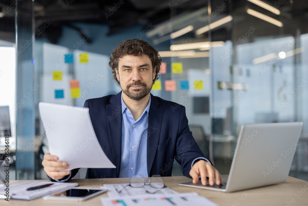 Serious businessman working in modern office holding document. Thoughtful professional analyzing information while using laptop. Focused on paperwork, enhancing productivity Serious businessman working in modern office holding document. Thoughtful professional analyzing information while using laptop. Focused on paperwork, enhancing productivity