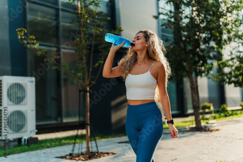 Canvas Print Fit young woman in blue leggings and a white crop top drinks water while walking, with trees and modern buildings in the background