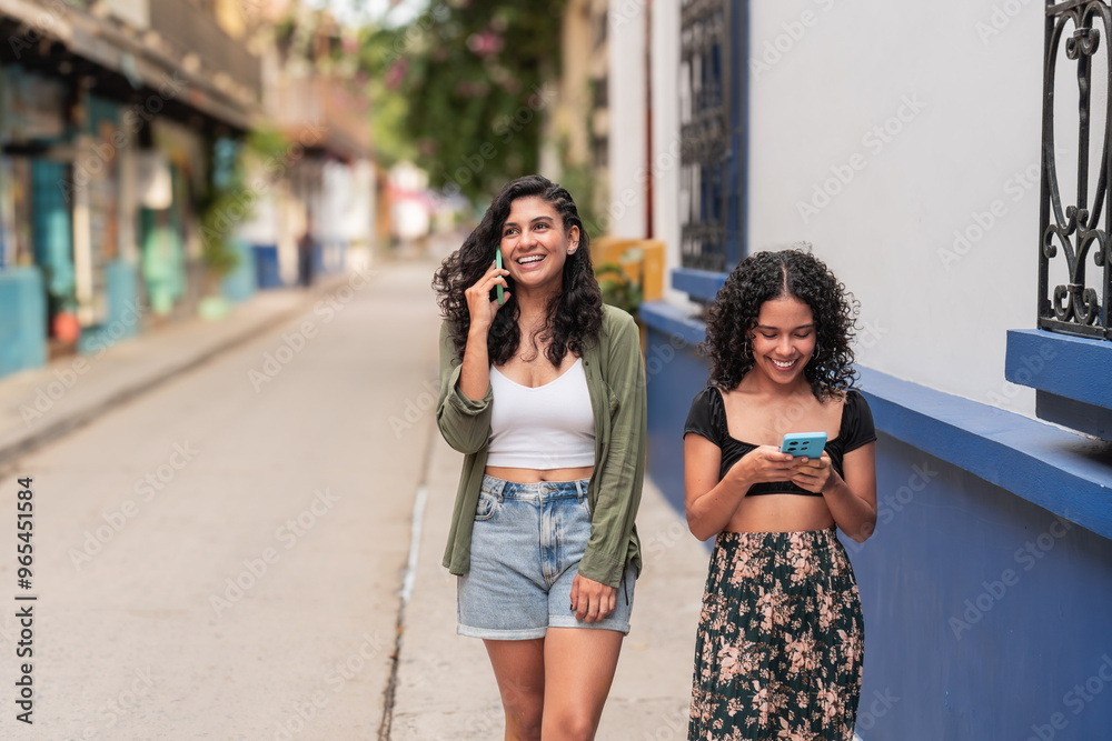 Two women walking outdoors while using smartphones on a sunny street. Two women walking outdoors while using smartphones on a sunny street.