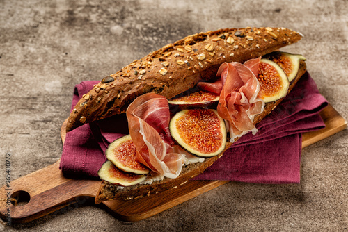 Sandwich with ham and figs. Seeded Baguette with Rye and Cereals, cheese, prosciutto crudo on a wooden board, with textile napkin, brown table background. photo