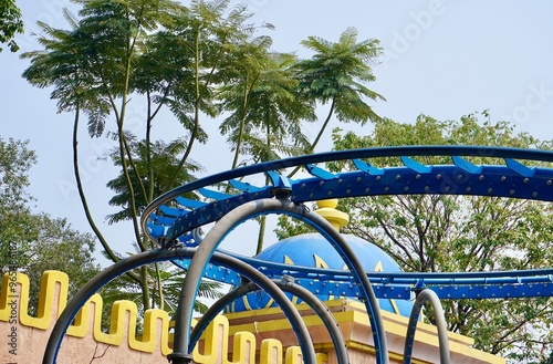 Blue roller coaster metal track rail way isolated on horizontal ratio outdoor sky, green botancal trees, and building exterior background. photo