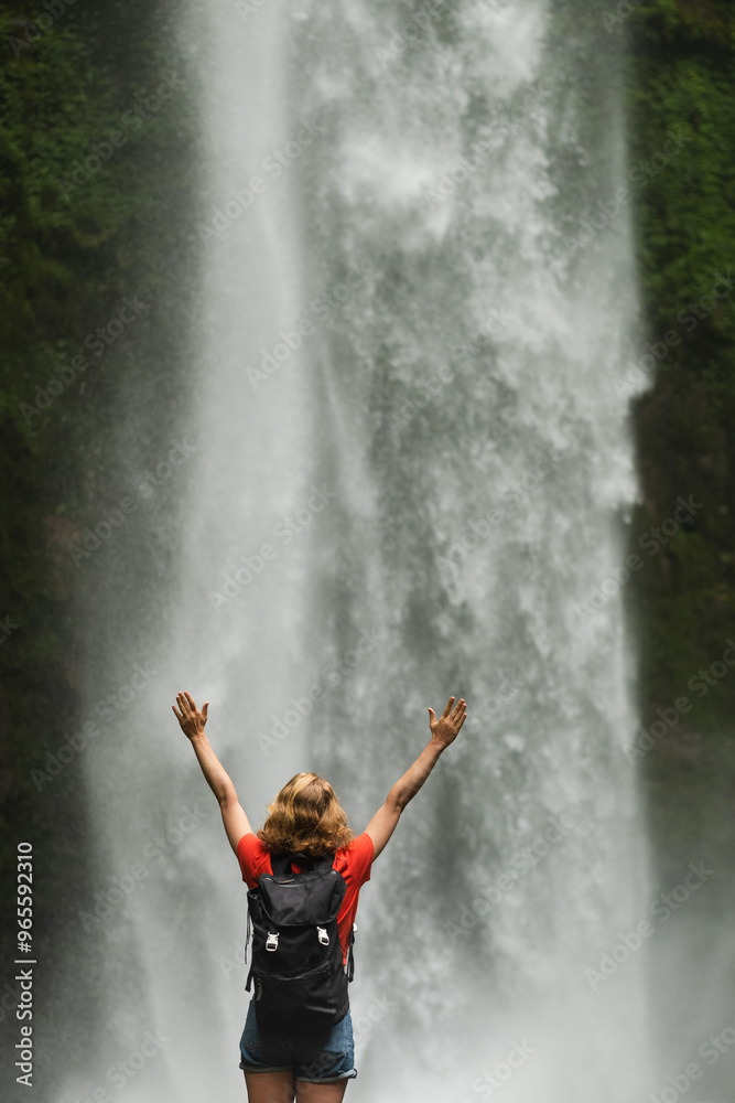 Obraz premium Blonde Woman Adventurer Standing in Front of Majestic Waterfall, Arms Raised in Triumph.