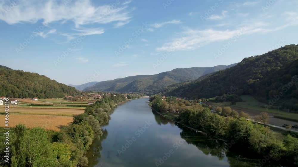 The Drina River flows serenely through a lush valley, surrounded by expansive green hills and cultivated farmland, under a clear blue sky with light clouds drifting above The Drina River flows serenely through a lush valley, surrounded by expansive green hills and cultivated farmland, under a clear blue sky with light clouds drifting above