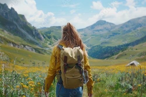 A young woman in a yellow jacket walks through a blooming valley surrounded by mountains on a sunny day during summer