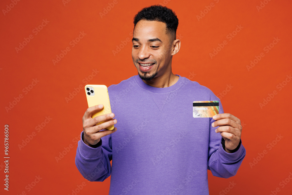 Young happy man of African American ethnicity wear purple sweatshirt casual clothes using mobile cell phone hold credit bank card shopping online booking tour isolated on plain red orange background. Young happy man of African American ethnicity wear purple sweatshirt casual clothes using mobile cell phone hold credit bank card shopping online booking tour isolated on plain red orange background.