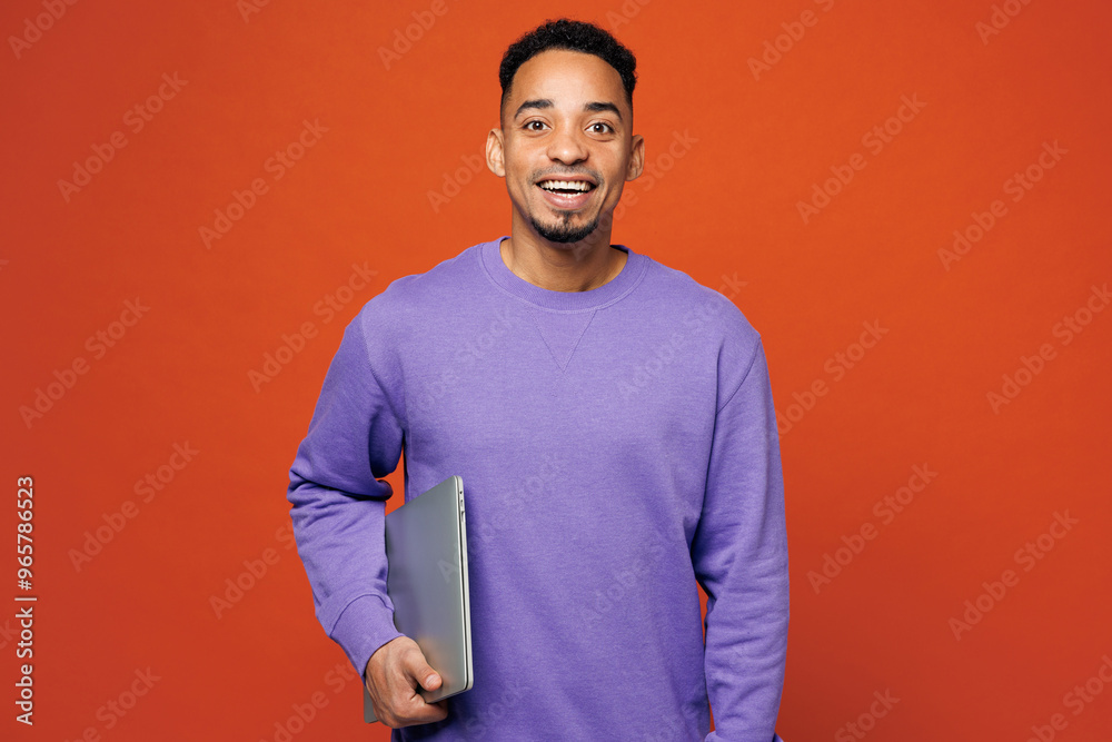 Φωτογραφία Young confident IT man of African American ethnicity he wear purple sweatshirt casual clothes hold closed laptop pc computer isolated on plain red orange background studio portrait