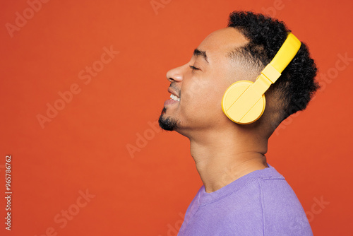 Close up side view young happy man of African American ethnicity wear purple sweatshirt casual clothes listen to music in headphones isolated on plain red orange background studio. Lifestyle concept. photo