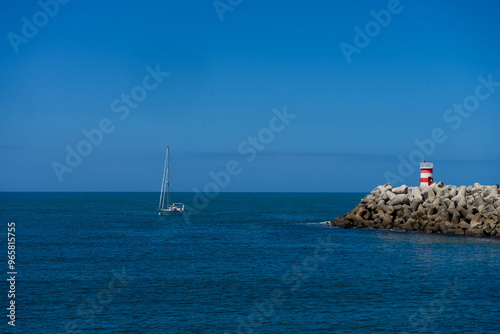 A boat approaches the entrance of Nazaré Marina, Portugal, with the lighthouse towers on both piers clearly visible, against a backdrop of calm waters and clear skies. photo