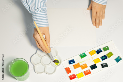 Artist mixing watercolor paints in ceramic palette in studio, close up. Female hands and watercolor accessories on table, top view