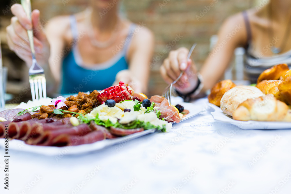 Friends enjoying a colorful spread of meats, salads, and bread at an outdoor gathering during a sunny day Friends enjoying a colorful spread of meats, salads, and bread at an outdoor gathering during a sunny day