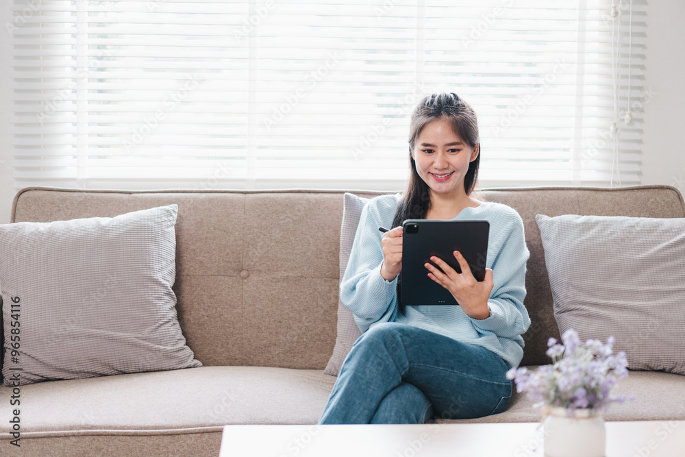 A young woman is sitting comfortably on sofa, using tablet while smiling. cozy living room features natural light and decorative cushions, creating warm atmosphere. A young woman is sitting comfortably on sofa, using tablet while smiling. cozy living room features natural light and decorative cushions, creating warm atmosphere.
