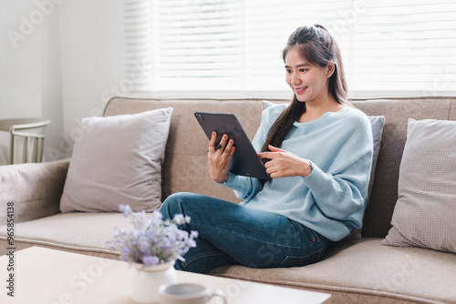 A young woman is sitting comfortably on sofa, using tablet while smiling. cozy living room features soft lighting and decorative cushions, creating warm atmosphere.