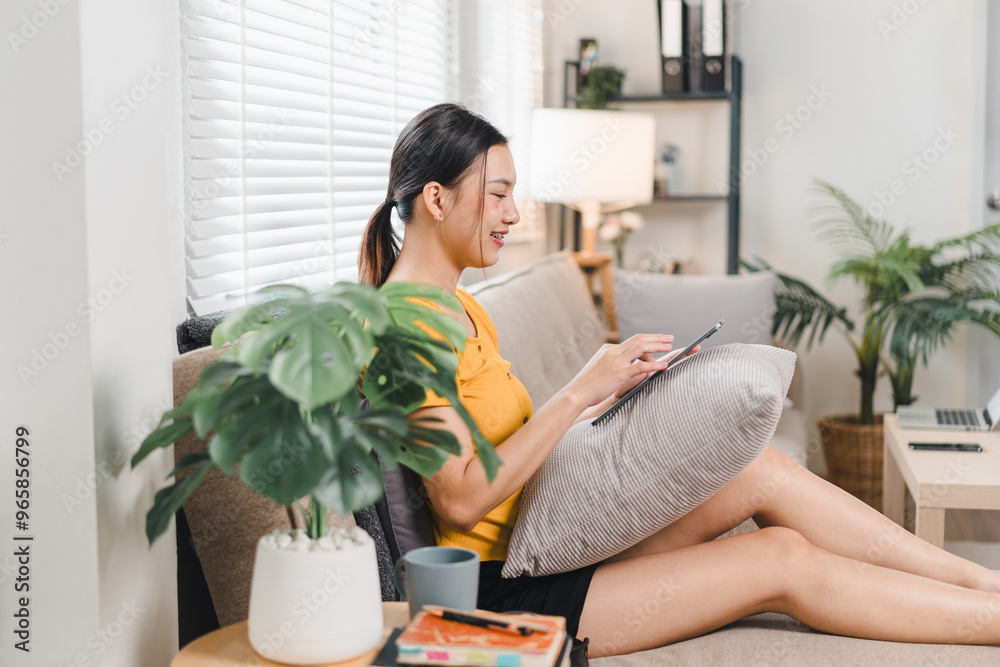 A young woman is sitting comfortably on sofa, using tablet while smiling. cozy living room features plants and natural light, creating relaxed atmosphere. A young woman is sitting comfortably on sofa, using tablet while smiling. cozy living room features plants and natural light, creating relaxed atmosphere.