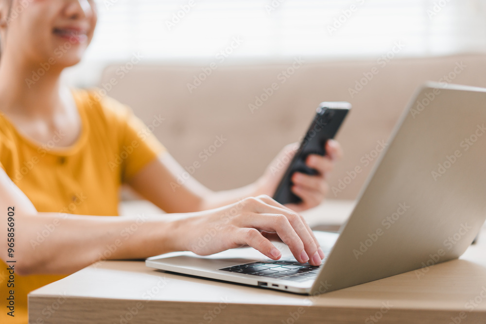 Fototapeta premium A woman in yellow shirt is using laptop while holding smartphone, showcasing modern workspace. scene reflects productivity and multitasking in cozy environment.
