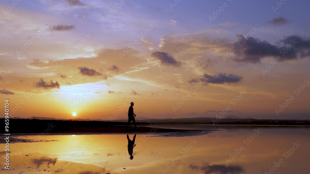 Man walks through shallow water at sunset, creating reflection in calm surface. As sun sinks below horizon, man becomes silhouette against colorful sky Man walks through shallow water at sunset, creating reflection in calm surface. As sun sinks below horizon, man becomes silhouette against colorful sky