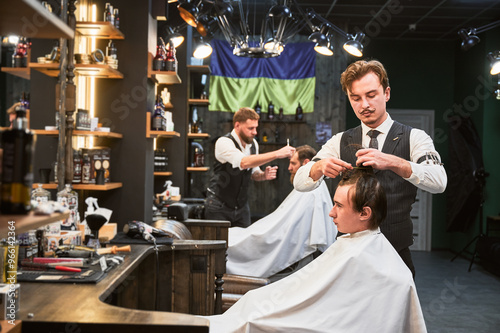 Professional barbers working on client hair in barbershop. Male hairstylists cutting hair in modern ambiance, with rustic wooden shelves stocked with various grooming products.