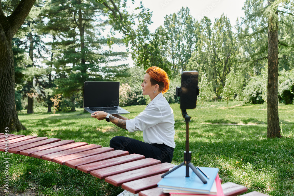 Fototapeta premium A teacher records a video outdoors, sitting on a bench in a park, showcasing her laptop.
