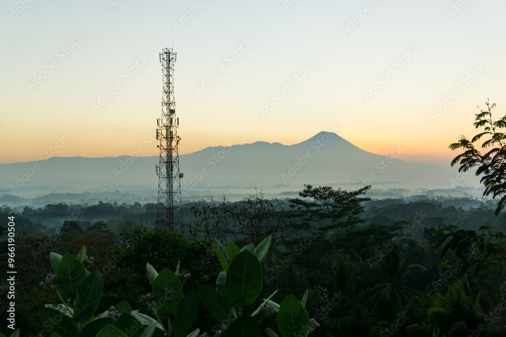 Obraz premium This image captures a peaceful morning landscape with a towering telecommunications tower set against the backdrop of a distant mountain at sunrise.