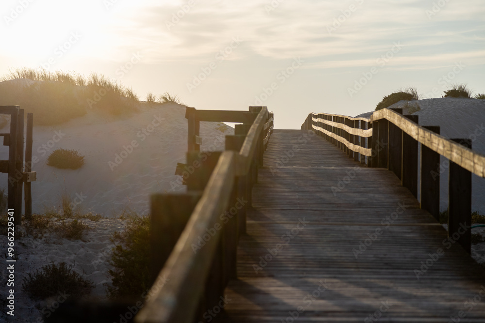 A wooden boardwalk between the sand dunes towards the sea at a beautiful sunset, with a soft golden light illuminating the path. Serene and perfect atmosphere on a peaceful walk at the end of the day. A wooden boardwalk between the sand dunes towards the sea at a beautiful sunset, with a soft golden light illuminating the path. Serene and perfect atmosphere on a peaceful walk at the end of the day.