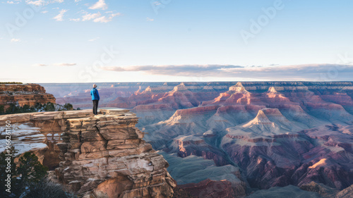 A photo of a tourist attraction, the Grand Canyon. There's a person standing on the edge of the canyon, looking out at the vast expanse below. The sky is clear, with a few clouds scattered across it. 