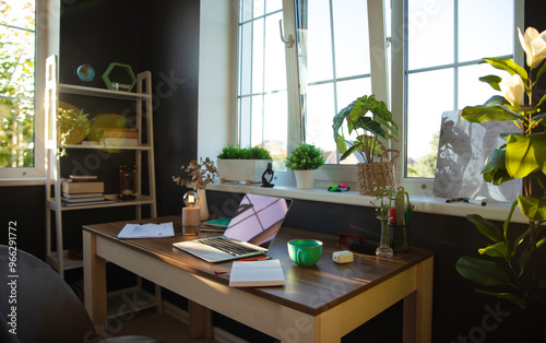 Modern, sunlit office desk is neatly arranged with laptop, documents, and coffee cup, surrounded by plants and personal items. Bright atmosphere and organized space reflect productivity and focus