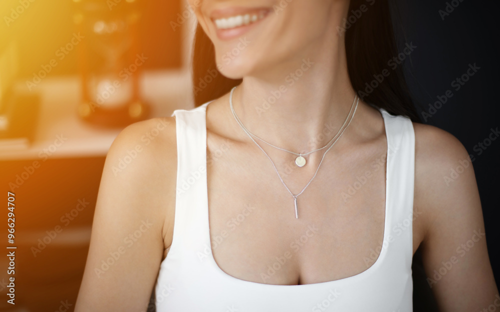 A close-up of a woman smiling while wearing two delicate silver necklaces, one with a small round pendant and the other with a vertical bar charm. A close-up of a woman smiling while wearing two delicate silver necklaces, one with a small round pendant and the other with a vertical bar charm.
