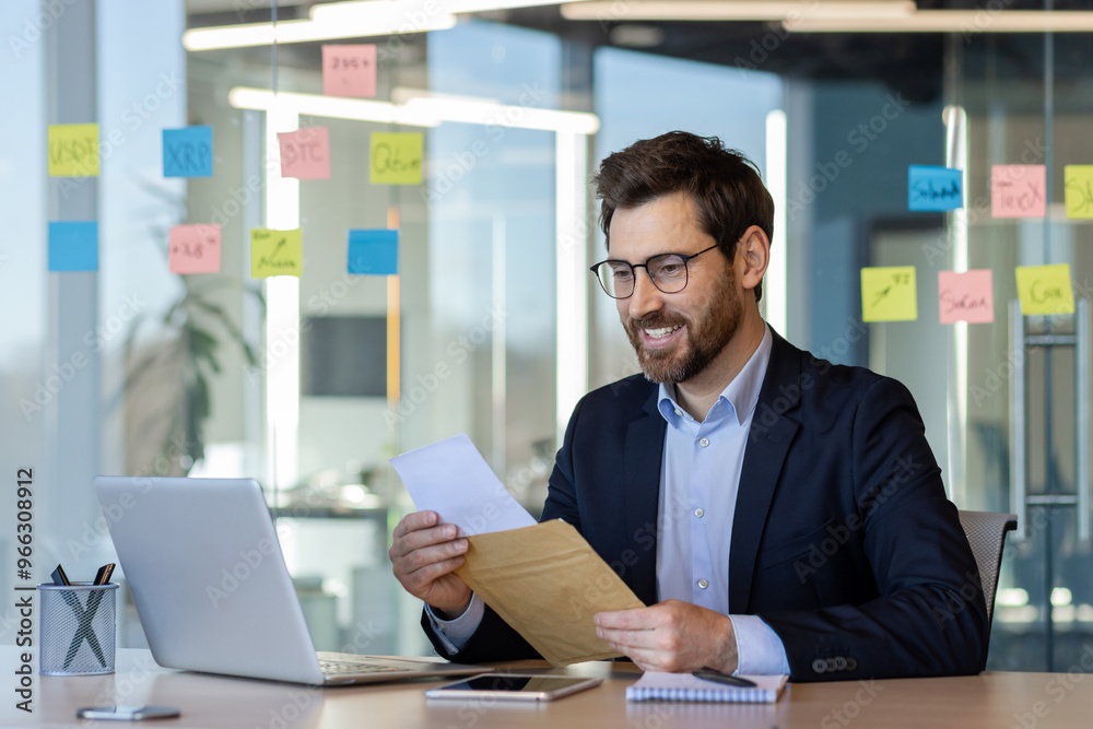 Business professional reading good news at desk in modern office. Smiling while holding document, seated with laptop and notepad. Concept of success, achievement, and workplace satisfaction.