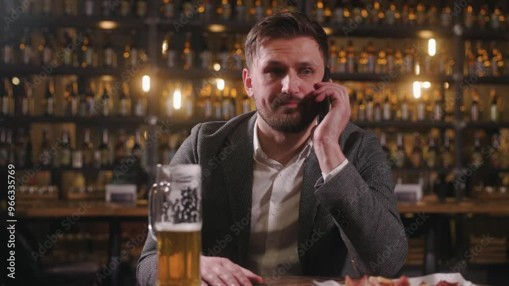 Man is sitting at bar table, engaged in a phone conversation while a half-full glass of beer and plate of food are in front of him. The background features dim, warm lighting with shelves of bottles. Man is sitting at bar table, engaged in a phone conversation while a half-full glass of beer and plate of food are in front of him. The background features dim, warm lighting with shelves of bottles.