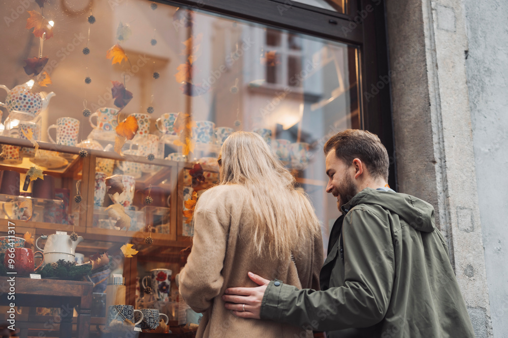 A couple admiring decorative pottery in a shop window. The woman has long hair and is wearing a beige coat, while the man in a green jacket stands close behind her, smiling. A couple admiring decorative pottery in a shop window. The woman has long hair and is wearing a beige coat, while the man in a green jacket stands close behind her, smiling.