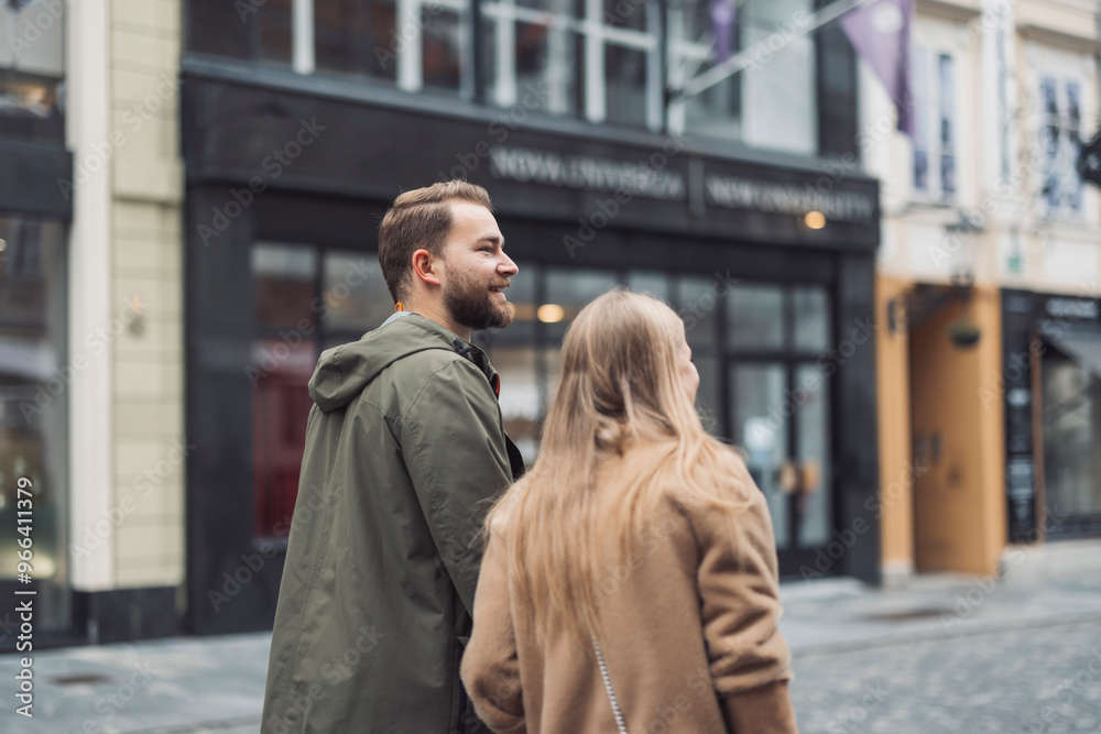 A couple walking together on a city street, smiling and enjoying their time. The man is wearing a green coat and the woman has long blonde hair, dressed in a beige coat. The background features shops A couple walking together on a city street, smiling and enjoying their time. The man is wearing a green coat and the woman has long blonde hair, dressed in a beige coat. The background features shops