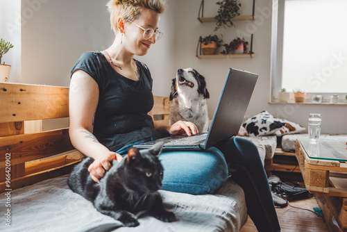 A woman sitting on a wooden sofa, working on a laptop. A black cat is resting on her lap, while a dog is playfully behind her. The room has plants and a cozy atmosphere. photo