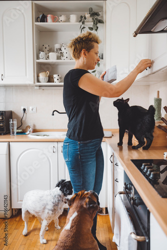 Canvas Print A woman in a black shirt and blue jeans stands in a kitchen, reaching for something on a shelf