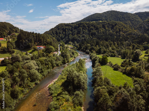 Aerial view of a serene river winding through lush green hills and forests, with scattered houses along the riverbank under a partly cloudy sky.