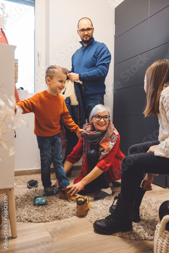 A family scene in a cozy hallway. A woman is helping a young boy put on his shoes while another adult stands nearby holding a coat. A girl sits on the floor, observing. The atmosphere is warm and invi photo