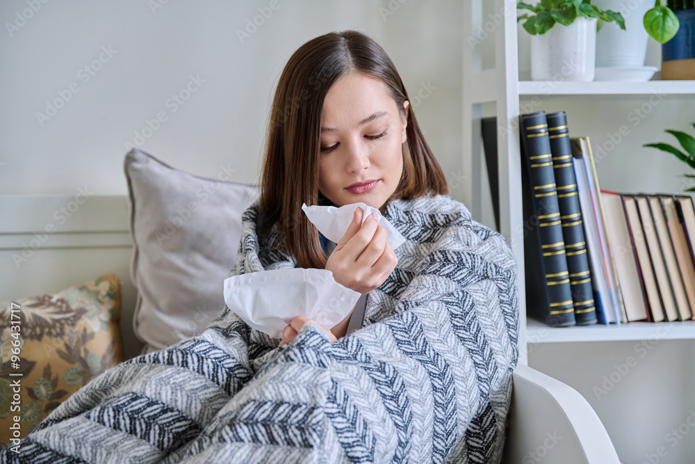 Young sick woman sitting under warm blanket sneezing coughing in handkerchief Young sick woman sitting under warm blanket sneezing coughing in handkerchief