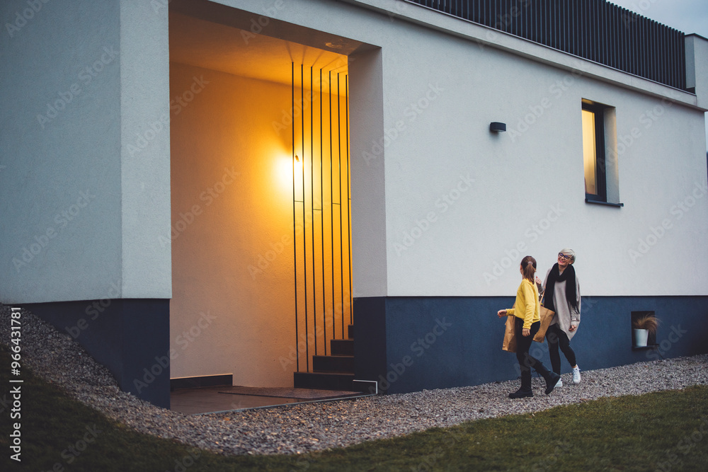 Fototapeta premium Mother and daughter coming home from a shopping together at dusk, with warm smart light illuminating the entrance. Enjoy spending time together. Holding bags with new clothes and shoes.