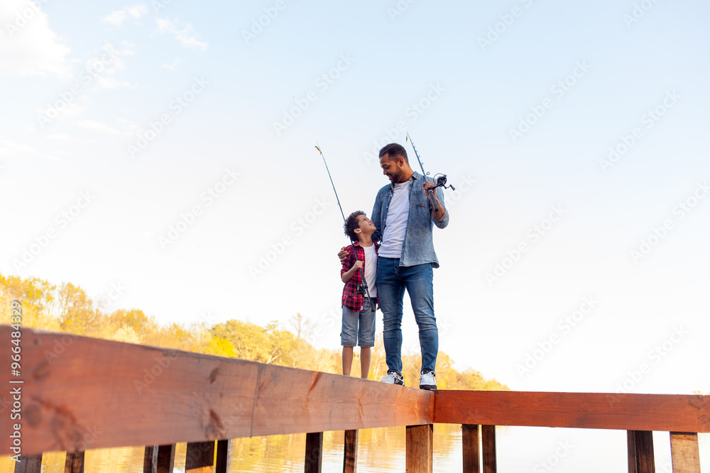 An African American father and his son stand on a wooden pier by a lake. The father, wearing a denim jacket and jeans, holds a fishing rod and hugs his son, looking at him with a smile. The son An African American father and his son stand on a wooden pier by a lake. The father, wearing a denim jacket and jeans, holds a fishing rod and hugs his son, looking at him with a smile. The son