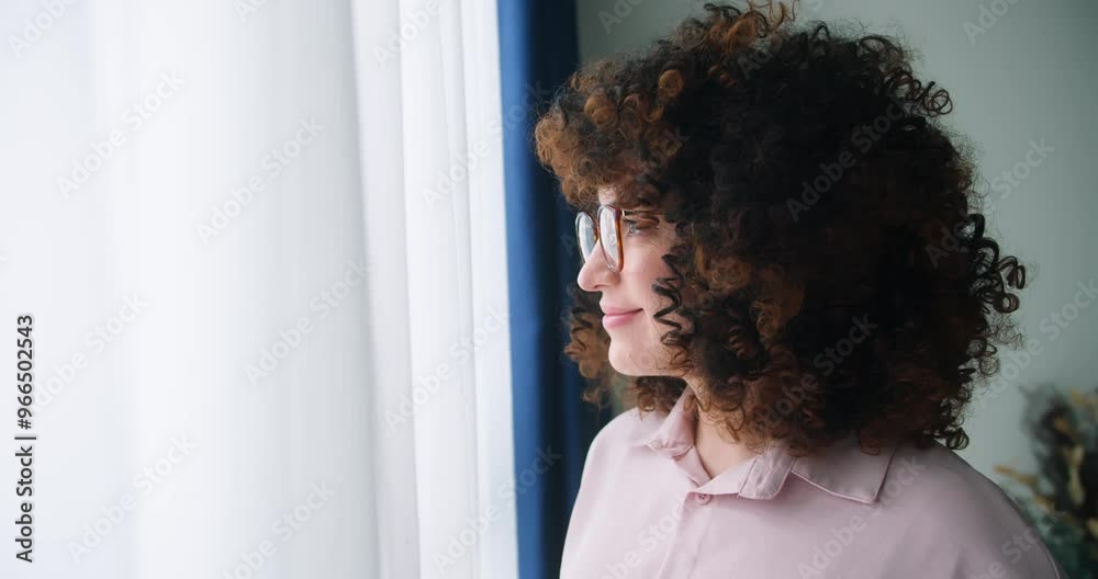 Curly girl looking out the window in her room. Woman with curly hair wearing glasses standing by the window. Curly girl looking out the window in her room. Woman with curly hair wearing glasses standing by the window.