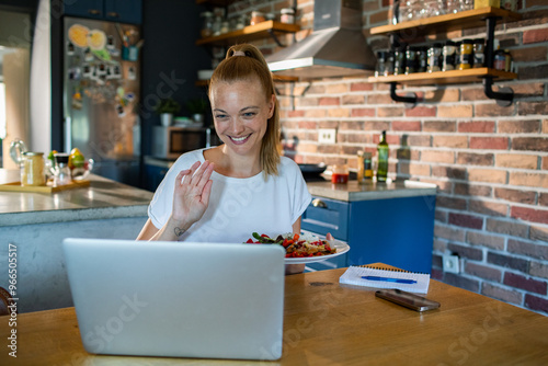 Smiling woman eating salad while video chatting from home kitchen