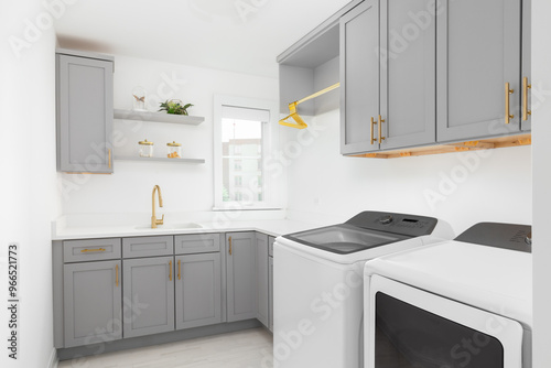 A laundry room with grey cabinets, white washer and dryer appliances, tile flooring, white countertop, and gold faucet and hardware. No brands or labels.
